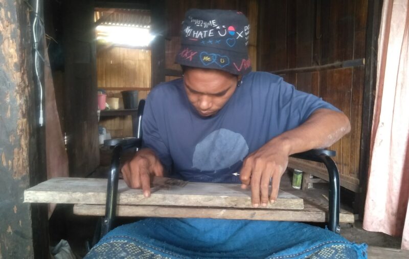 Reza sedang melukis nama di atas papan kayu untuk selanjutnya di pahatnya walaupun di atas kursi roda (Foto: Chen Rasi/Savanaparadise.com)